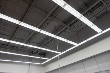 White commercial strip lights hanging from an exposed ceiling of pipes and beams in a large retail store