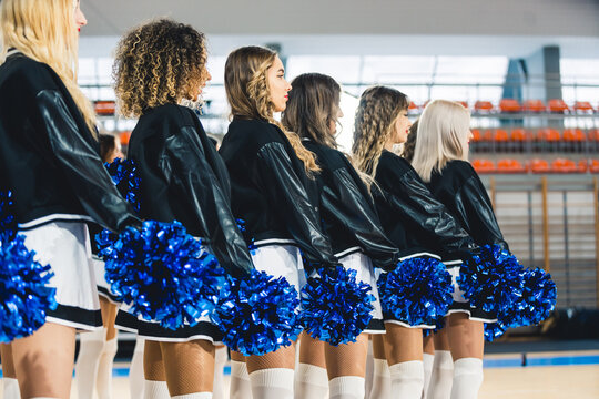 Side view of cheerleaders in uniforms standing in line with blue pom-poms in their hands. Blurred sports hall in the background. High quality photo