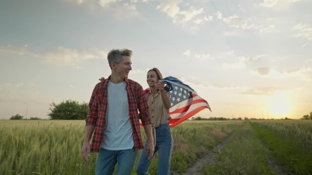4k slow motion of happy patriotic couple celebrating fourth of july with national flag in rural lardmark at sunset. 4th of July. USA independence day celebrating with American flag.