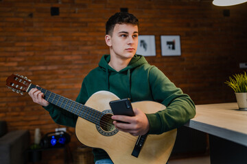 One young man Caucasian teenager sit at home in room playing guitar use smartphone mobile phone to...