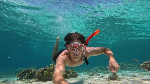 A man in a red snorkel mask dives underwater, swimming next to the white sand at the bottom of the sea. A man on vacation in the Maldives dives under warm sea water. Exploring seabed by amateur diver.