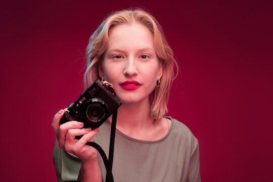 A happy blonde photographer is holding camera while standing in front of the magenta background. Viva magenta.