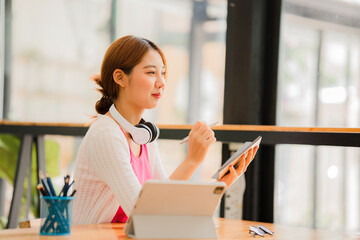 Asian female college student with headphones on her neck enjoying online classes Watching videos and taking notes from digital tablet homework, online learning concept