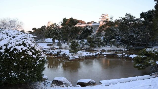 Dawn light hits Japanese castle tower overlooking pond in snow covered park
