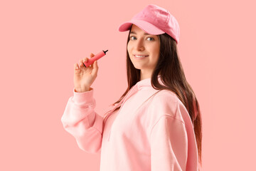 Young woman in cap with disposable electronic cigarette on pink background