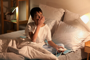 Fototapeta premium Sleepy little boy sitting on electric heating pad in bedroom at night