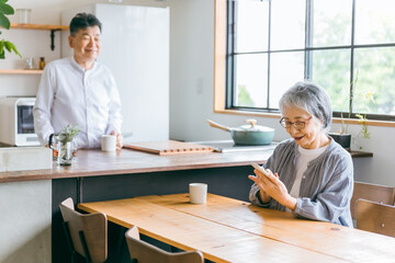 家のダイニングで笑顔でスマホを使う高齢者女性と男性(老夫婦)