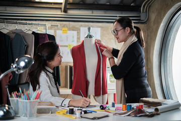 Asian middle-aged female fashion designer teaches young teen trainee tailor about shape and size with measure tape and sewing fabric for dress design collection, professional boutique small business.