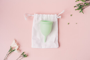 Green menstrual cup on white pouch next to dry flowers on pink background.