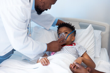 Fototapeta premium African american male doctor giving ventilator to boy patient with mother in hospital and copy space