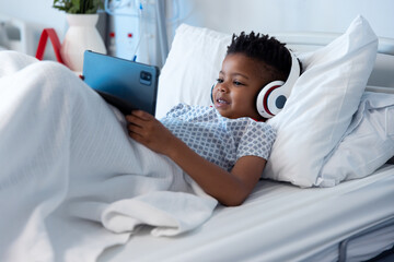 Happy african american boy patient in headphones using tablet in hospital bed