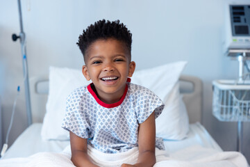 Happy african american boy patient sitting up in hospital bed, smiling with copy space