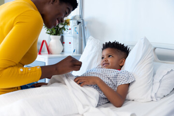 Happy african american mother sitting with son patient in hospital bed, smiling with copy space