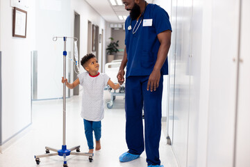 Happy african american male doctor holding hand of boy patient with drip in corridor with copy space