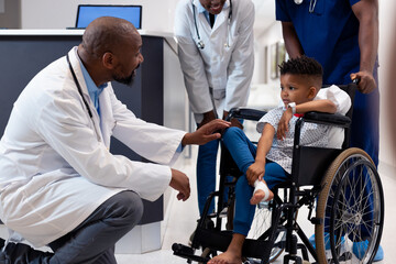 Fototapeta na wymiar Smiling african american male doctor talking to boy patient in wheelchair in corridor