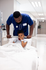 Vertical of african american male doctor pushing boy patient in hospital bed in corridor