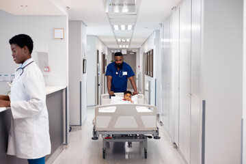 African american male doctor pushing boy patient in hospital bed in busy corridor with copy space