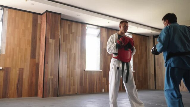 Two martial arts men in kimonos and gloves practice kicks and punches. Male fighters wearing kimono, gloves and protection belts training sparring at gym. Kick boxers jumping and practicing actions