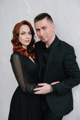 Stylish beautiful photo session in a photo studio of a luxurious couple in a black suit and black dress, where a girl with red hair. They hug.