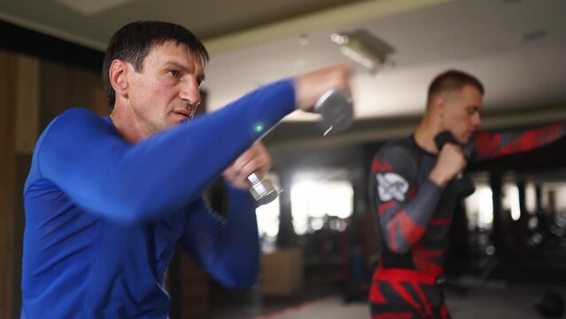 Male boxers training activities with dumbbells at fitness club. Male fighters exhausting box training kicks with dumbbells. Adult and fit men having evening workout indoor. Fitness and sport concept
