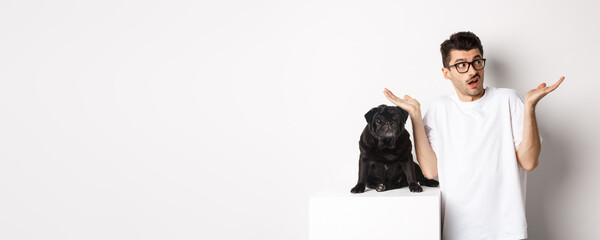 Portrait of confused hipster guy dog owner shrugging, standing near cute black pug pet, white...