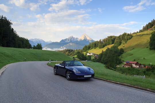 Berchtesgaden, Germany - July 25, 2021: Blue roadster Porsche Boxster 986 with mountain Watzmann and fog panorama. The car is a mid-engine two-seater sports car manufactured by Porsche.