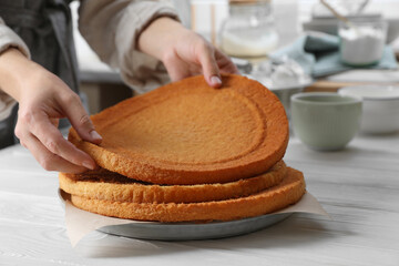 Woman stacking homemade sponge cakes at white wooden table in kitchen, closeup