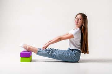 A teenage girl in jeans and a white t-shirt stretches her legs with green and purple styrofoam cubes. White background. The concept of sports, yoga.