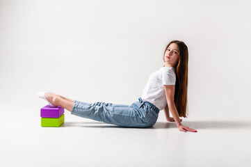 A teenage girl in jeans and a white t-shirt stretches her legs with green and purple styrofoam cubes. White background. The concept of sports, yoga.