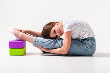 A teenage girl in jeans and a white t-shirt stretches her legs with green and purple styrofoam cubes. White background. The concept of sports, yoga.