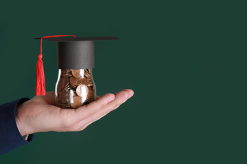 Man holding glass jar of coins and graduation cap against green background, closeup with space for text. Scholarship concept