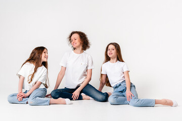 Mom and two teenager daughters in jeans and white t-shirts are smiling and happy. They sit with their knees bent. White background.