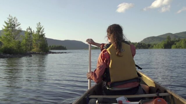 Female paddles a canoe on a lake with mountains in the background during sunset in slow motion