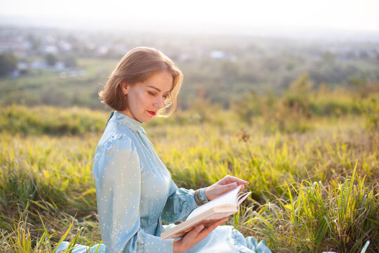 A Woman In A Long Summer Dress With Short Hair Sitting On A White Blanket And Reading The Book. Concept Of Having Picnic In A City Park During Summer Holidays Or Weekends. 