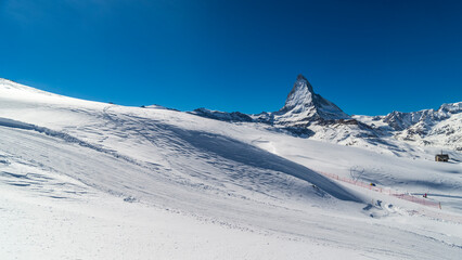 Fototapeta premium Ski slope in Swiss Alps in sunny day,Matterhorn behind. Small wooden house near by ski slope.Mountains and skiing resort for winter travel.Winter landscape of Switzerland.Zermatt winter landscape.