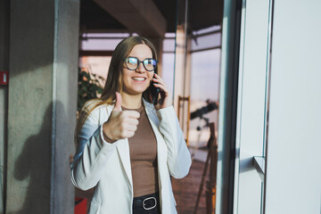 A fair-haired European woman in a white jacket and glasses is talking on the phone while standing in a spacious office with large windows. Successful young woman freelancer