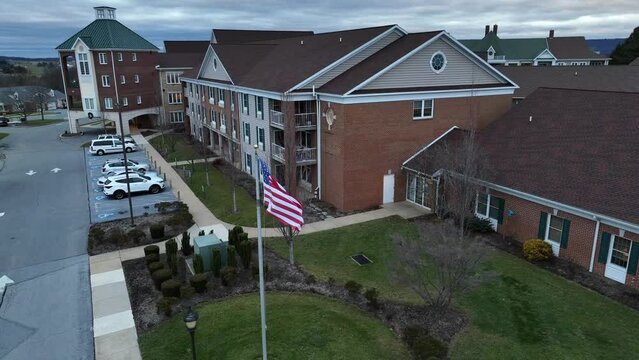 Aerial Rising Shot Of American Flag Waving In Front Of Brick Retirement Home Building. Assisted Living In America Theme.