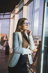 A pretty woman in glasses is standing in the office near a large window. Successful businesswoman in her office. Business portrait