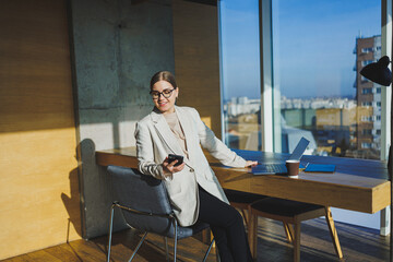 Cute positive female employee with long blond hair in casual clothes looking at phone while working on new business project at table with laptop and gadgets in office