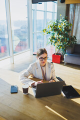 A positive business woman in a jacket is sitting at a table, working on a laptop in a bright office. A young European woman is sitting at a table. Freelance and remote work. Modern women's lifestyle