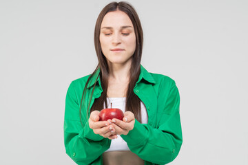 A cute brown-haired girl holds a red ripe apple in her hands on a white background. The model is wearing a green shirt and blue jeans.