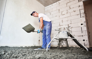 Male worker using shovel while shoveling sand-cement mix in building under construction. Man preparing floor screed material while standing near concrete screed mixer machine.