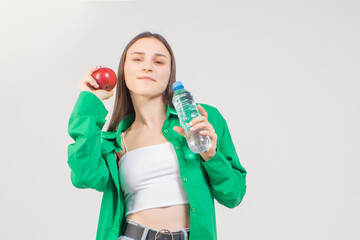 A young pretty brown-haired girl holds a small bottle of clean water and a red apple. The model is wearing a green shirt, white top and jeans, she is smiling.