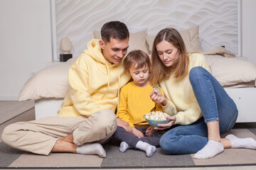 Cute family, parents in yellow clothes with child son sitting by the bed and eating popcorn