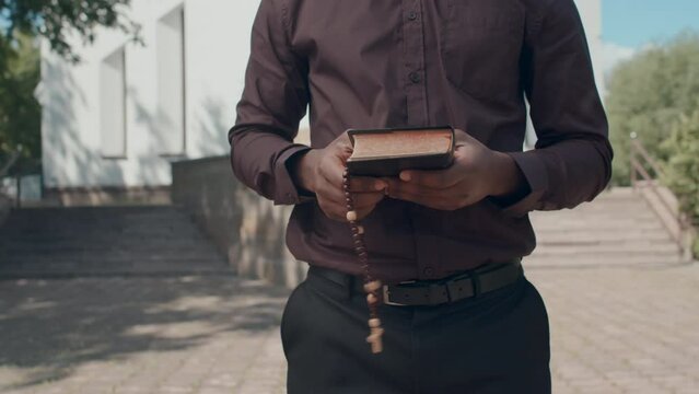 Medium section of unrecognizable Black man holding prayer beads standing outdoors reading the Bible