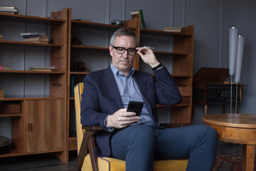 Handsome elderly grey haired successful businessman in glasses looking on his mobile phone, sitting in office