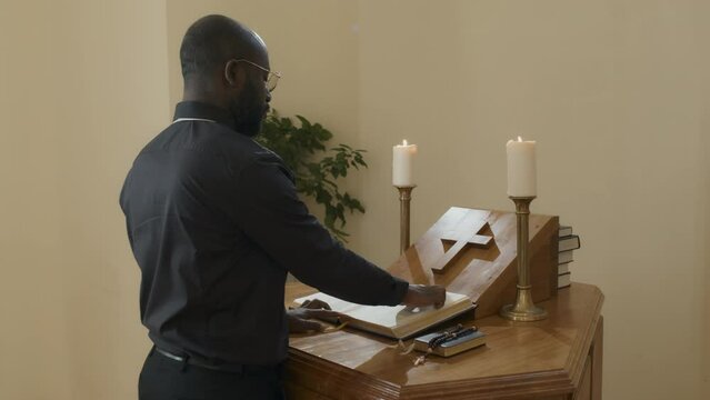 Medium shot of young adult African American priest standing at wooden altar in Catholic church reading the Bible