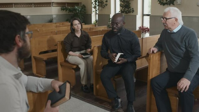 Modern young adult African American Catholic pastor sitting on bench in church explaining Bible story to parishioners