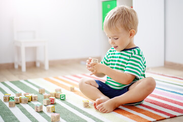 Cute boy sitting on the carpet indoor and playing with wooden cubes.