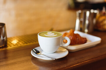 March 13, 2018, Belarus, Minsk. Horizontal photo of a white illy coffee cup with matcha cappucсino and a plate with a croisant on a wooden bar counter.